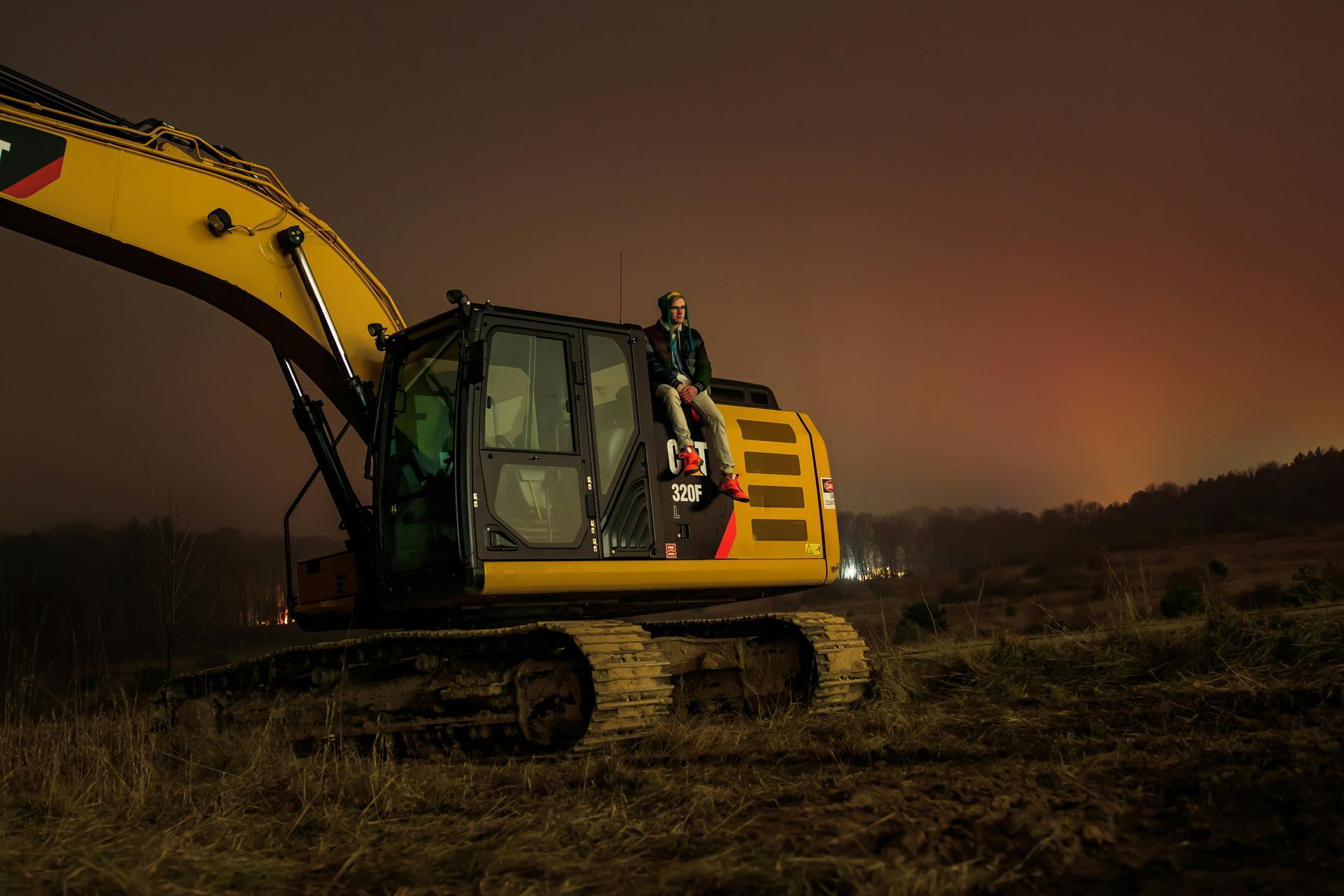 A person sits on a yellow excavator cab in a dark field at night.