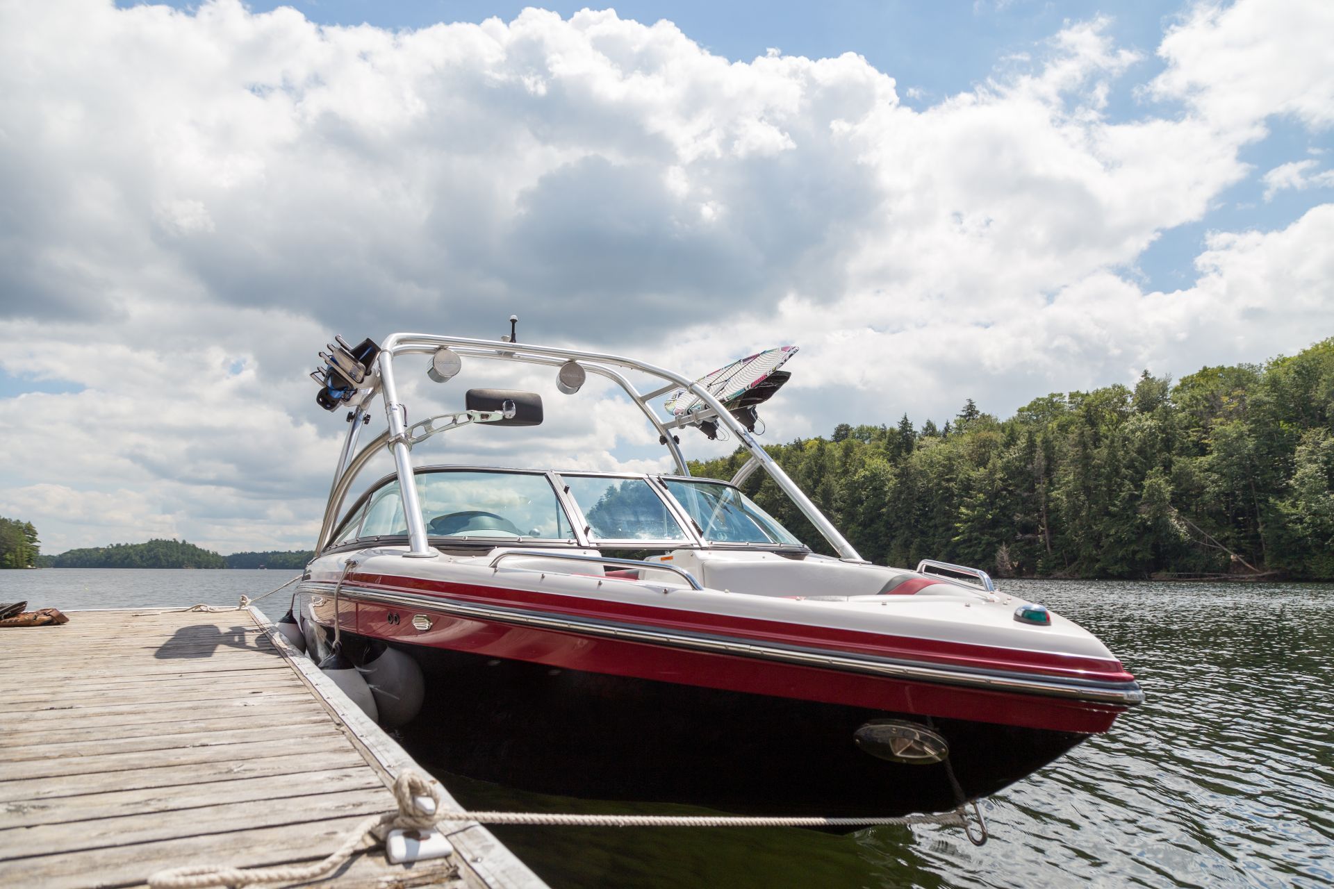 Boat docked on a lake, black and red hull, white windshield, silver frame, cloudy sky, trees in background.