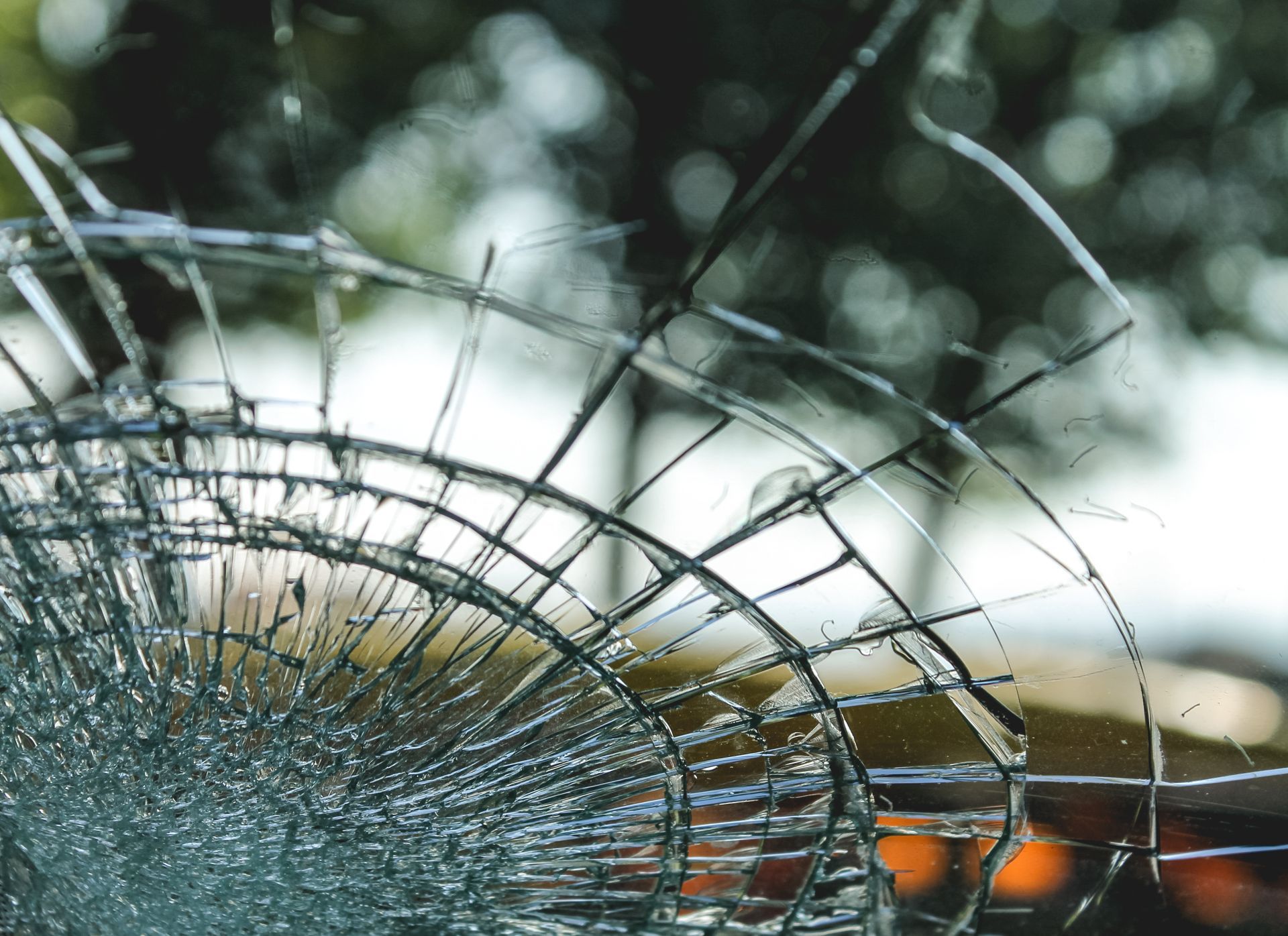 Cracked car windshield with a radial pattern against a blurred background of trees and light.