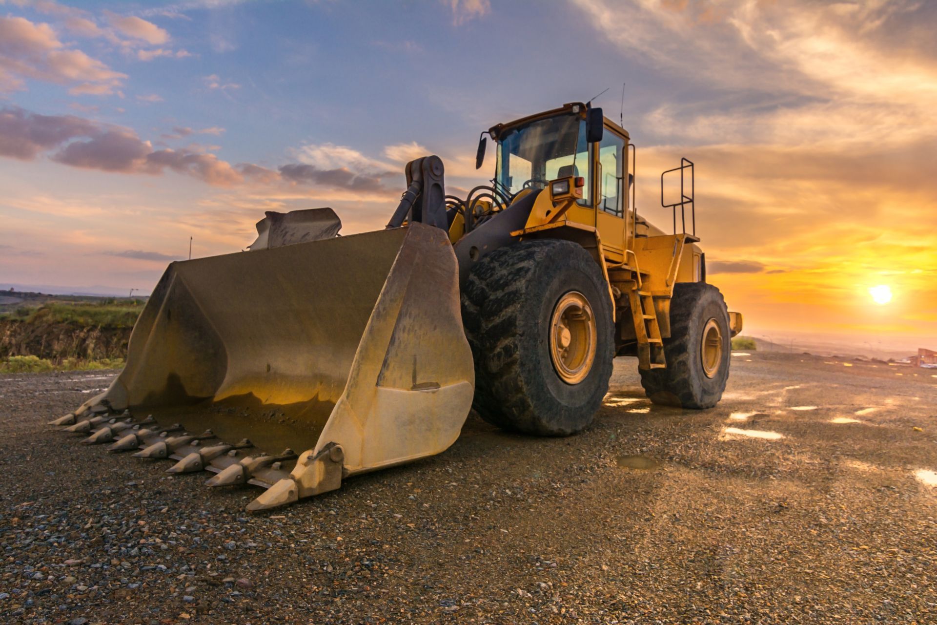 Yellow front-end loader on gravel with sunset in background.