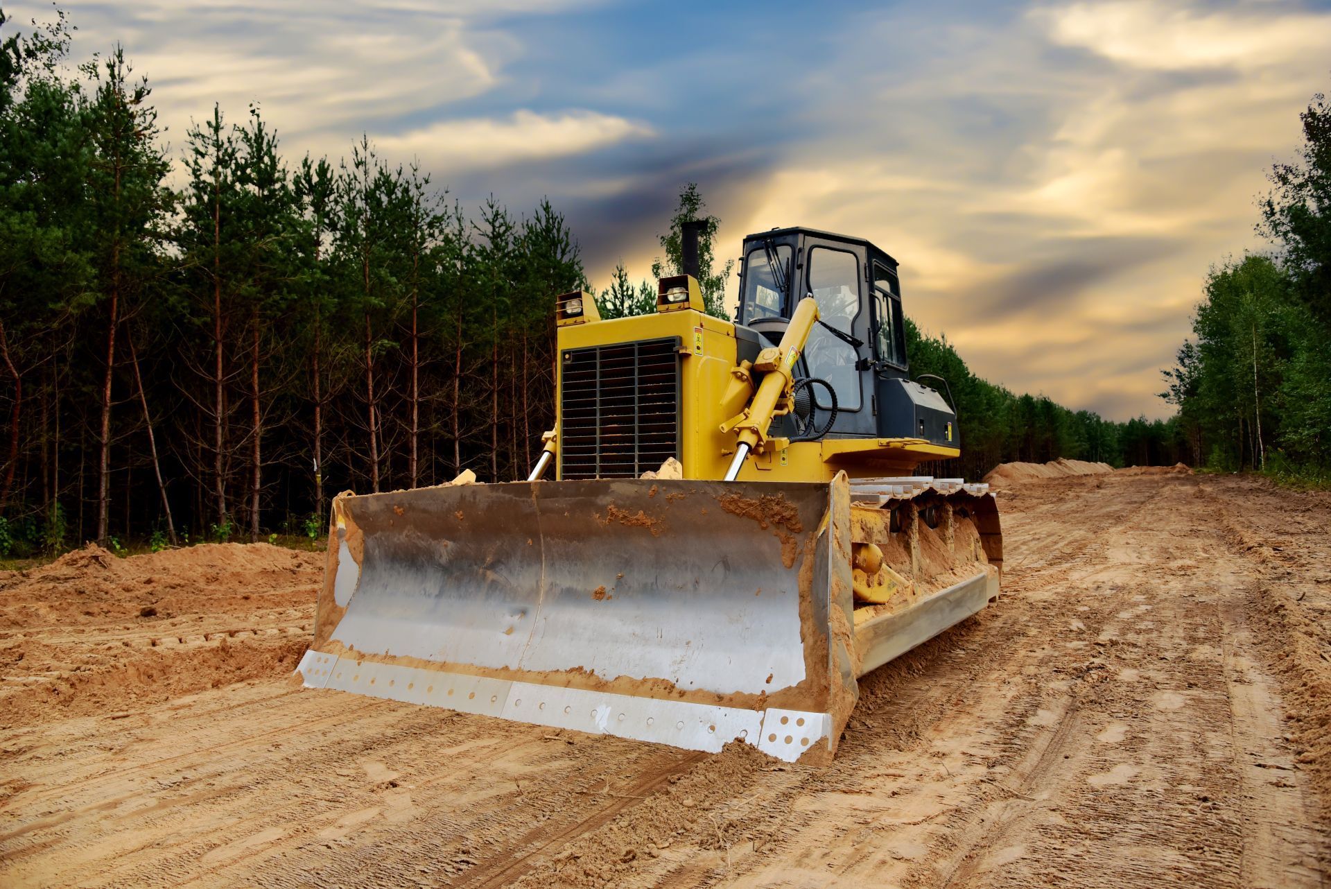 Yellow bulldozer on dirt road, clearing land near a forest. Cloudy sky overhead.