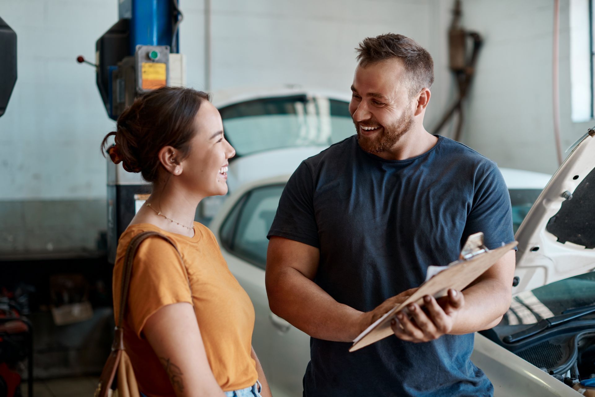 Woman smiles at auto mechanic in garage, discussing paperwork near a car with an open hood.