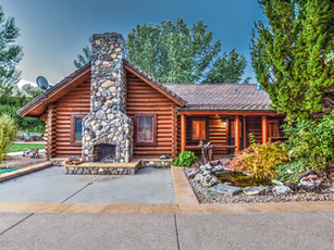 Rustic log cabin with stone chimney, front porch, and landscaped yard under a blue sky