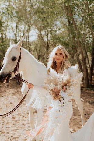 Blonde woman in a white dress standing beside a white horse in a sunny wooded setting