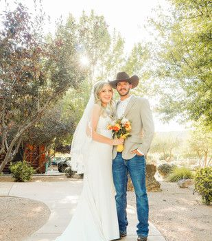 Bride and groom posing outdoors in bright sunlight, holding a bouquet, with trees and a path behind them