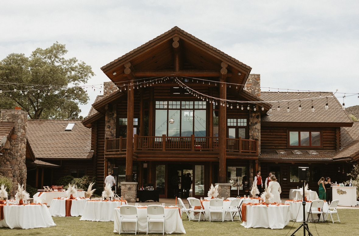 Outdoor wedding reception setup in front of a rustic wooden lodge, with white tablecloth tables and chairs on the lawn.