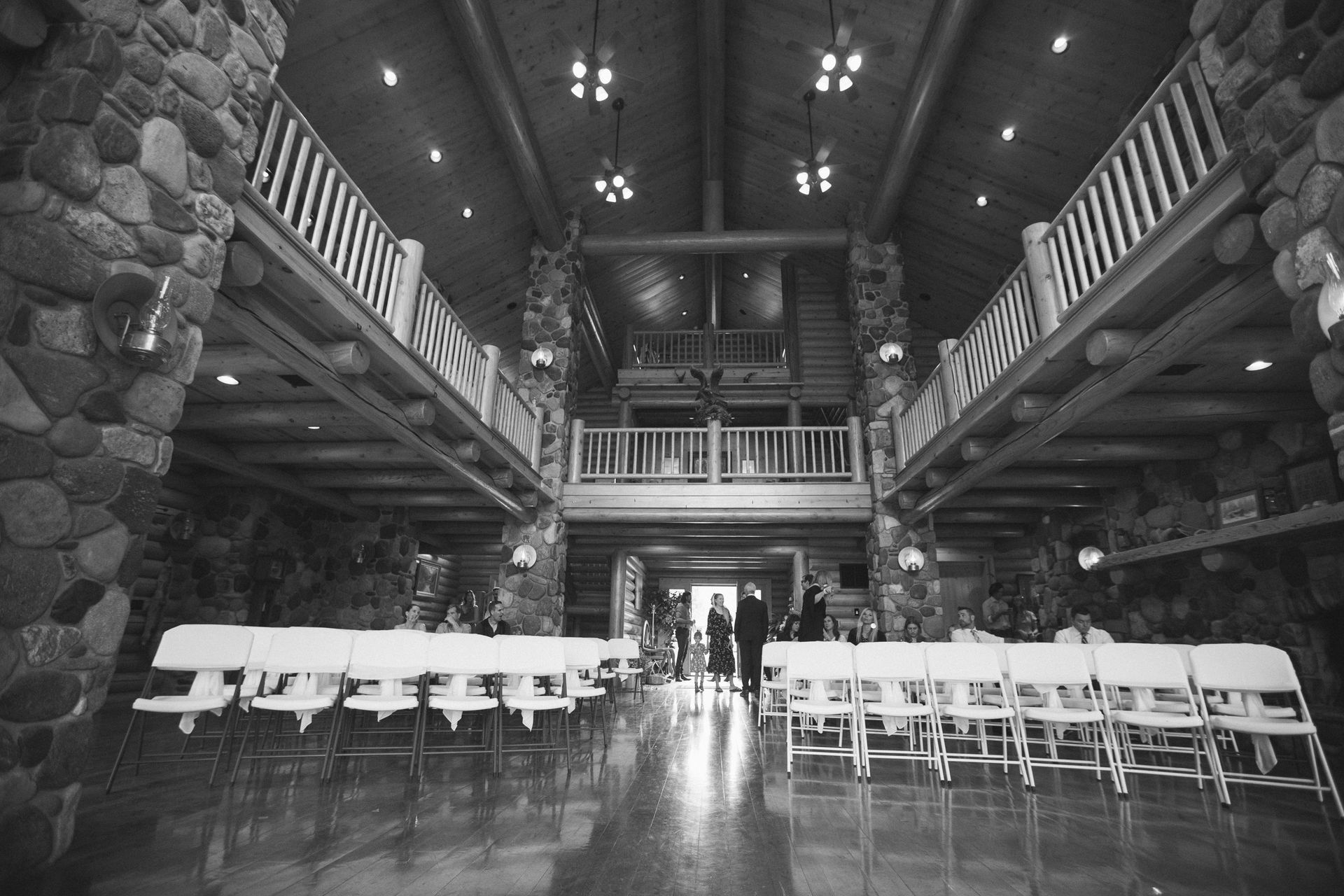 Wide lodge-style banquet hall with long tables, white chairs, stone walls, and a high wooden ceiling