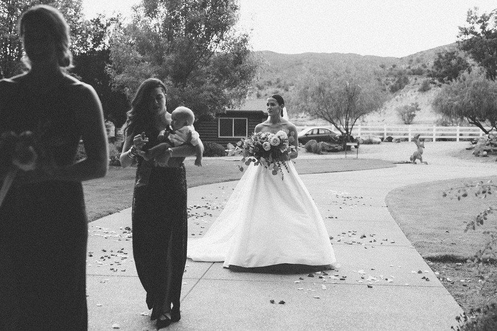Bride in white dress holding bouquet on a path at an outdoor wedding, with guests in the foreground