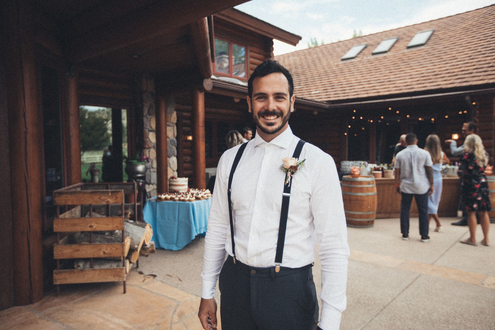 Smiling man in white shirt and suspenders stands outside a rustic building at a wedding venue