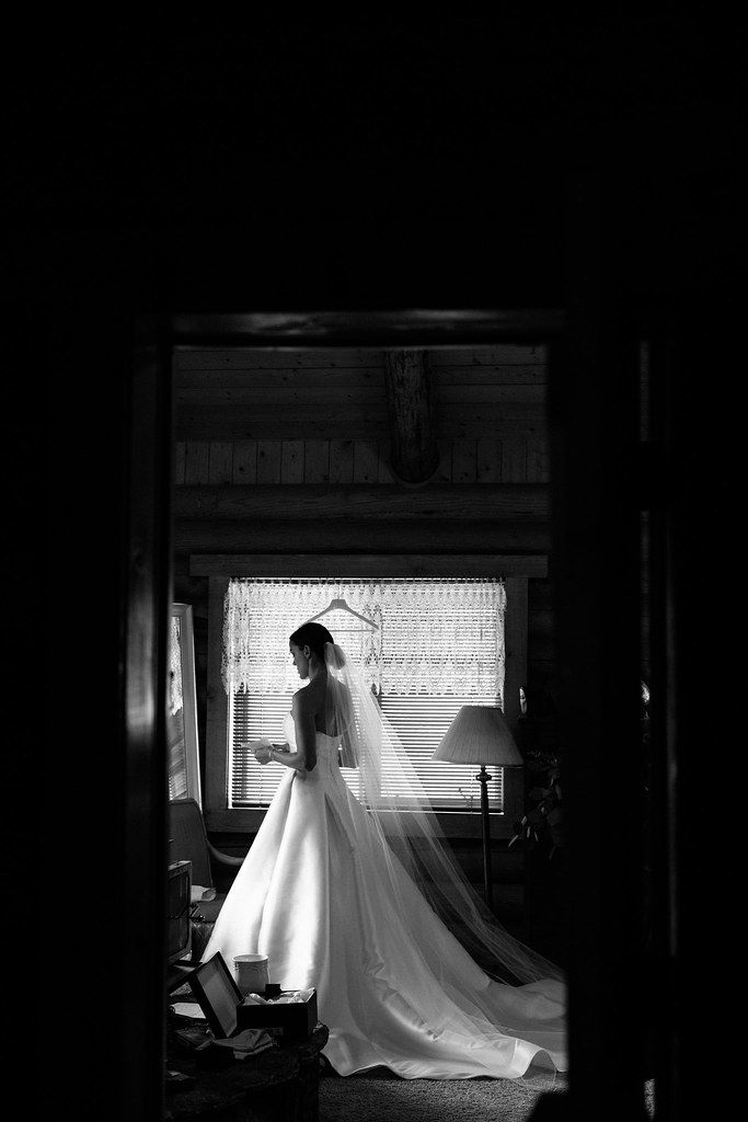 Bride in a long white gown standing in a doorway, backlit in a dark room.