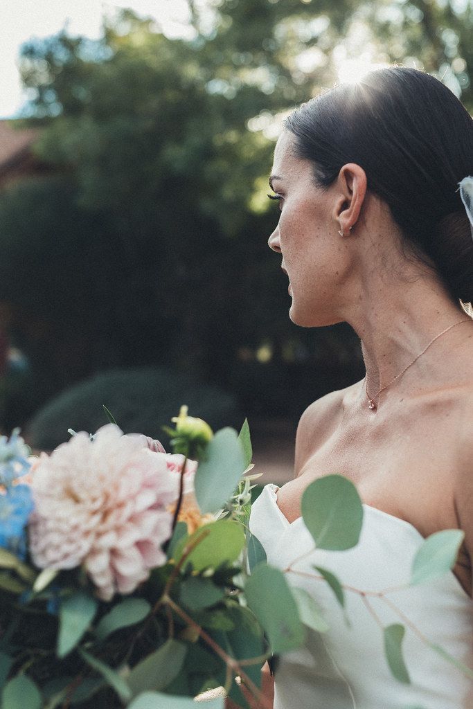 Woman in a white dress holding a bouquet, looking to the side outdoors with trees in the background