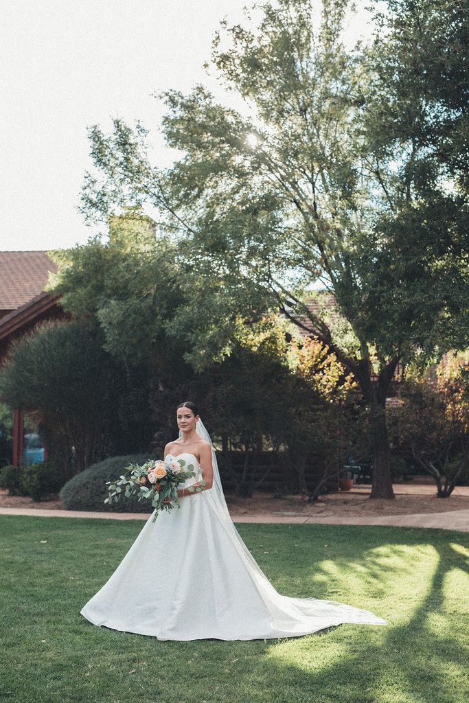 Bride in a white gown holding a bouquet in a grassy garden with trees and a building behind her.