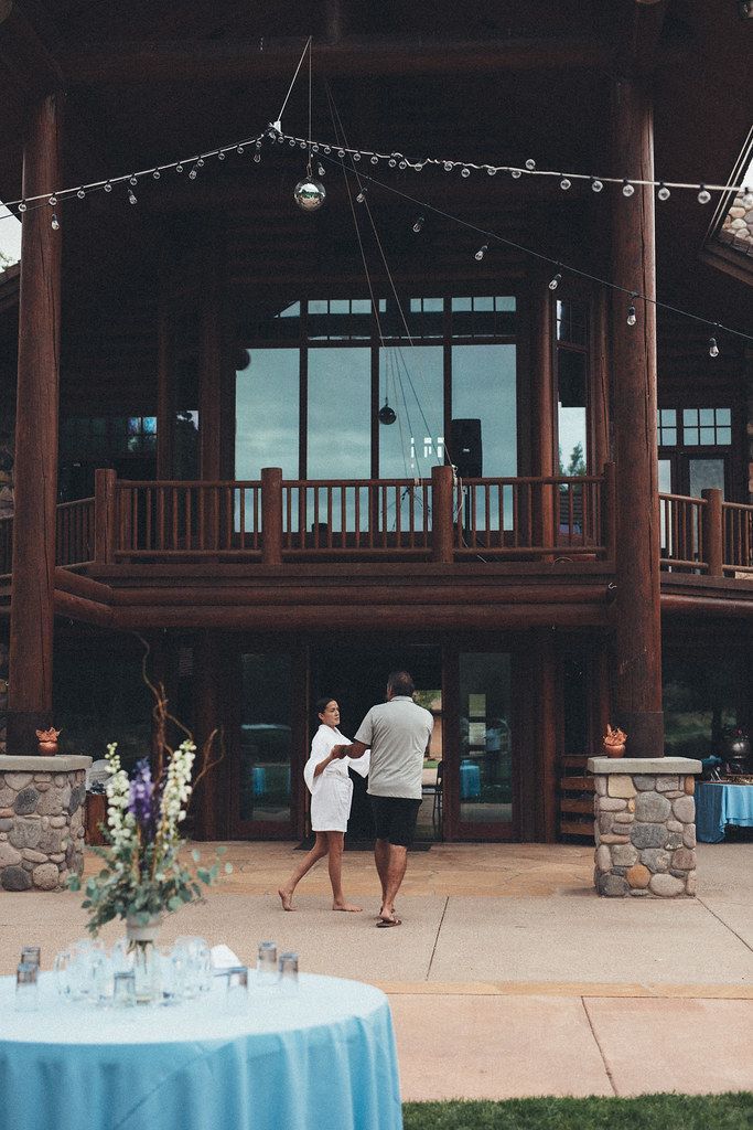 Two people walk toward a rustic lodge entrance with stone pillars and a blue fountain in front.