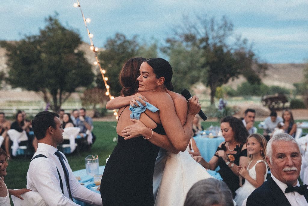 Two people embrace at an outdoor wedding reception, with guests seated at tables in the background.
