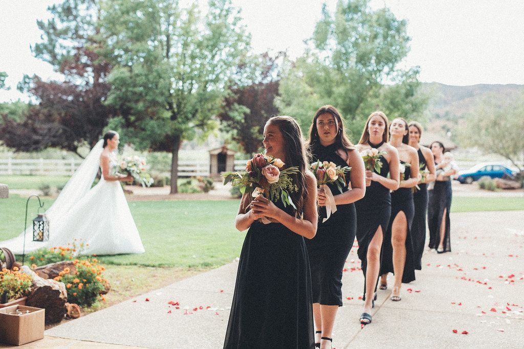 Bride in white stands beside a line of bridesmaids in black dresses outdoors at a wedding ceremony.