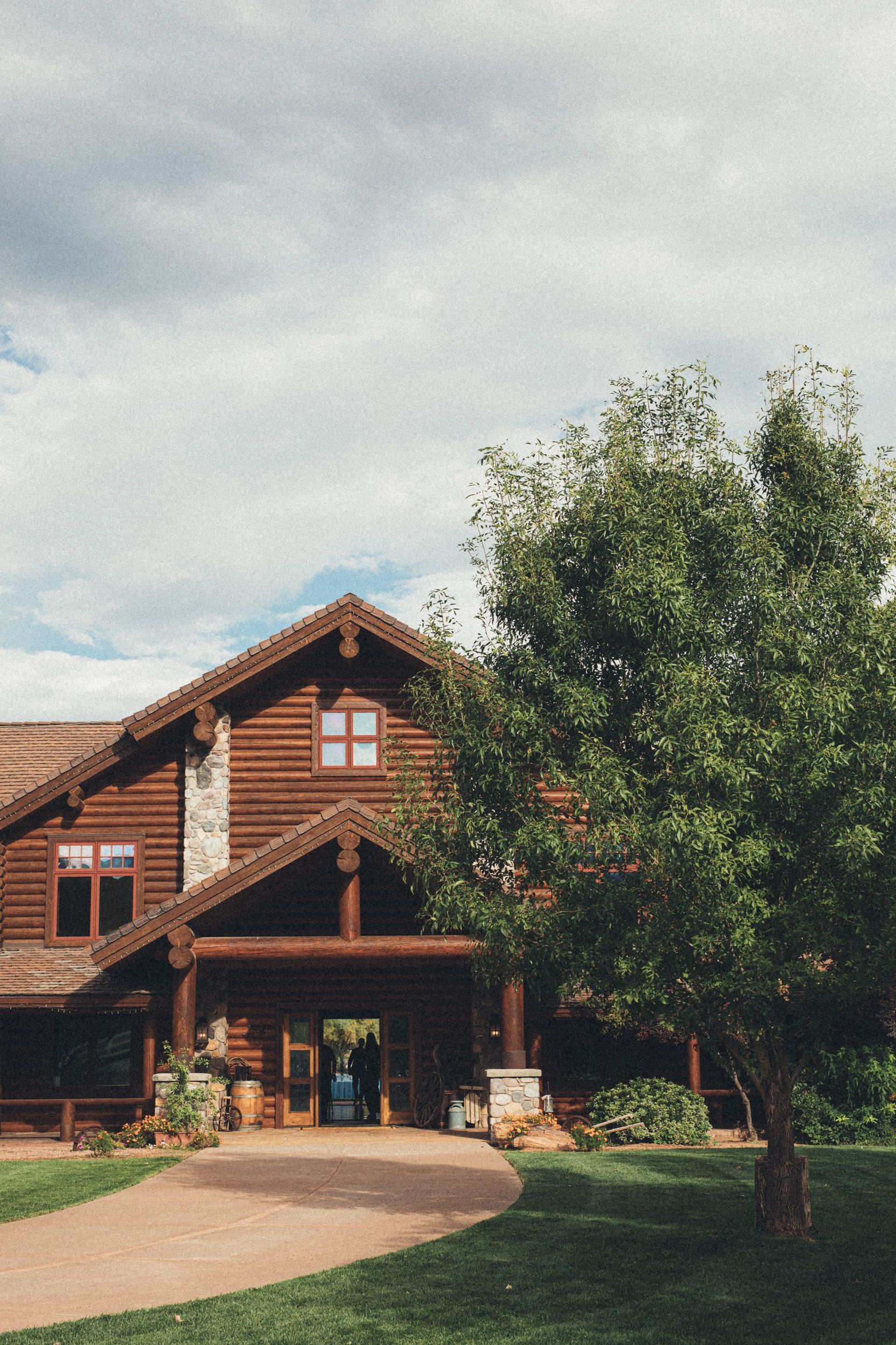 A rustic wooden house with a porch, a large tree in front, and a cloudy sky.