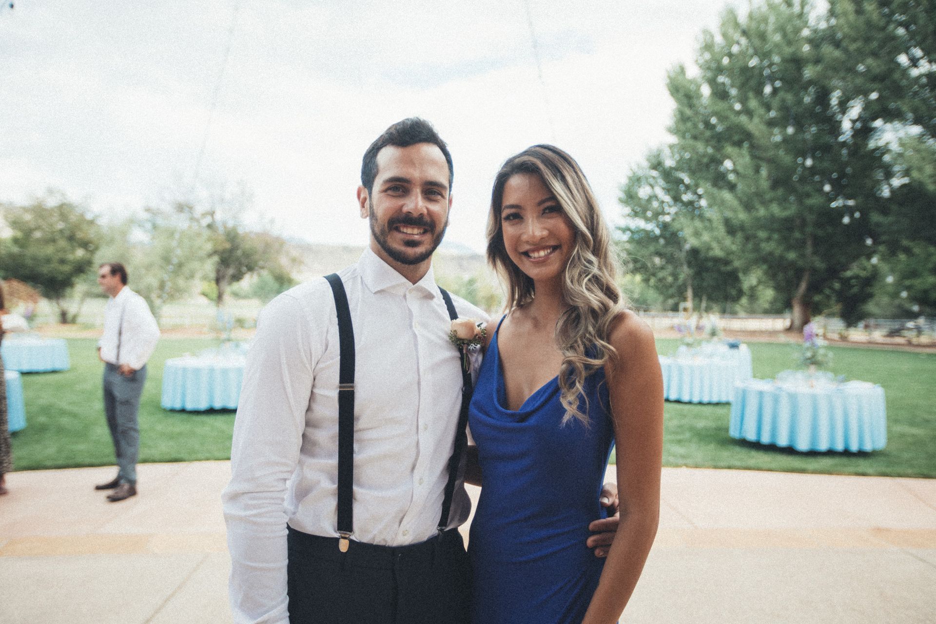 Smiling couple posing outdoors at an event, with blue table settings and trees in the background.