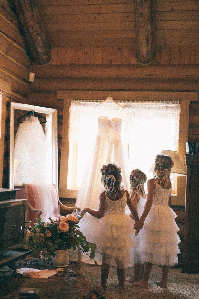 Two young children in white dresses standing by a bright window in a rustic room