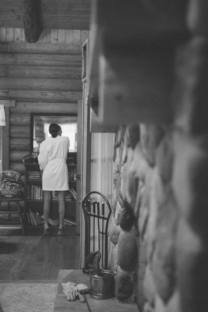 Person in a white coat stands in a rustic hallway, facing away near a doorway.