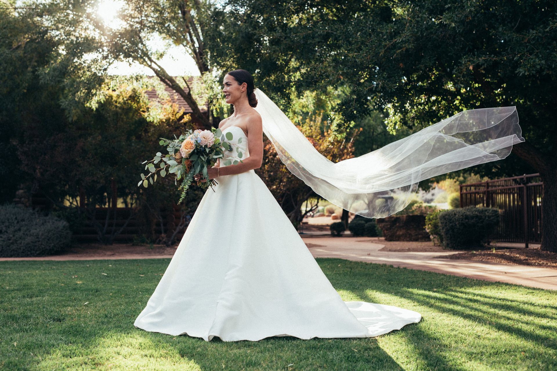 Bride in a white gown holding a bouquet outdoors, with a long veil flowing behind her.
