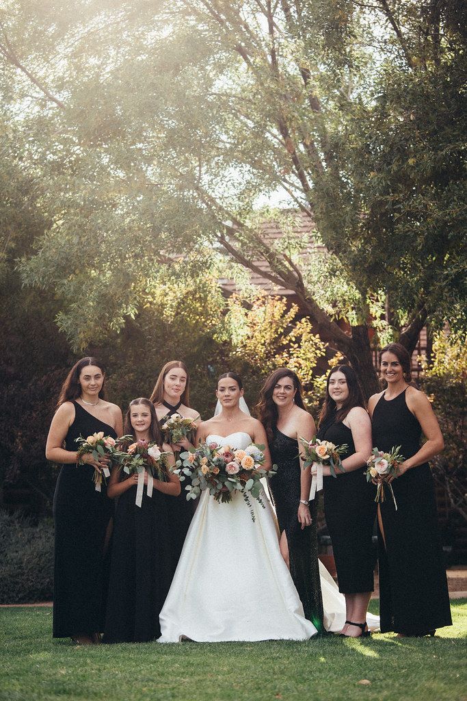 Wedding party posing outdoors in black dresses and a white gown, holding bouquets under trees.