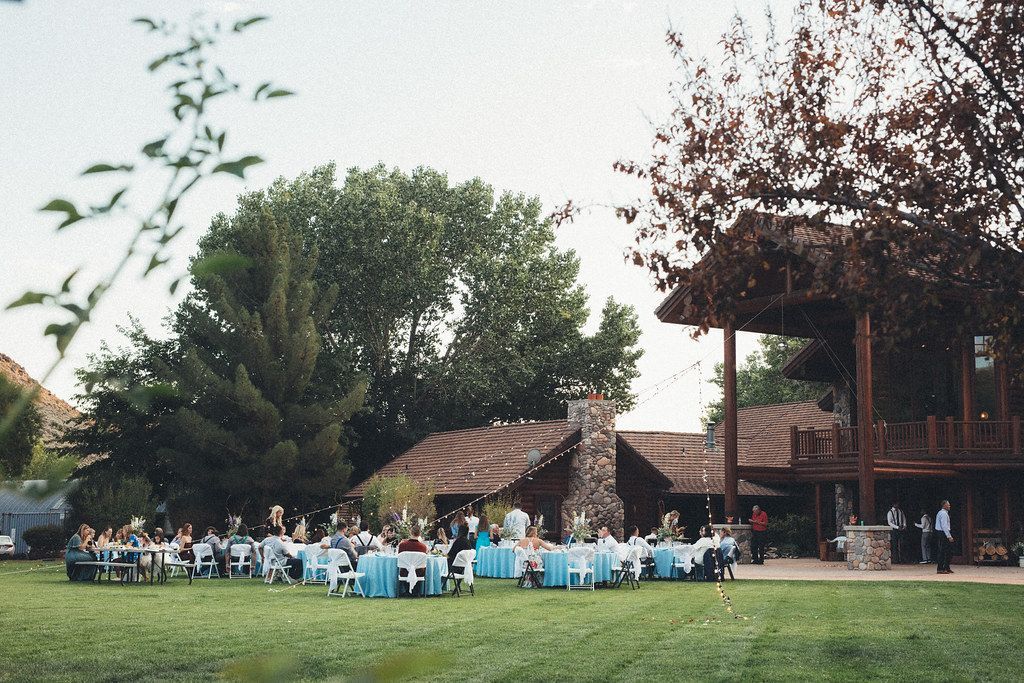 People seated at blue-clothed tables on a lawn beside rustic buildings and trees