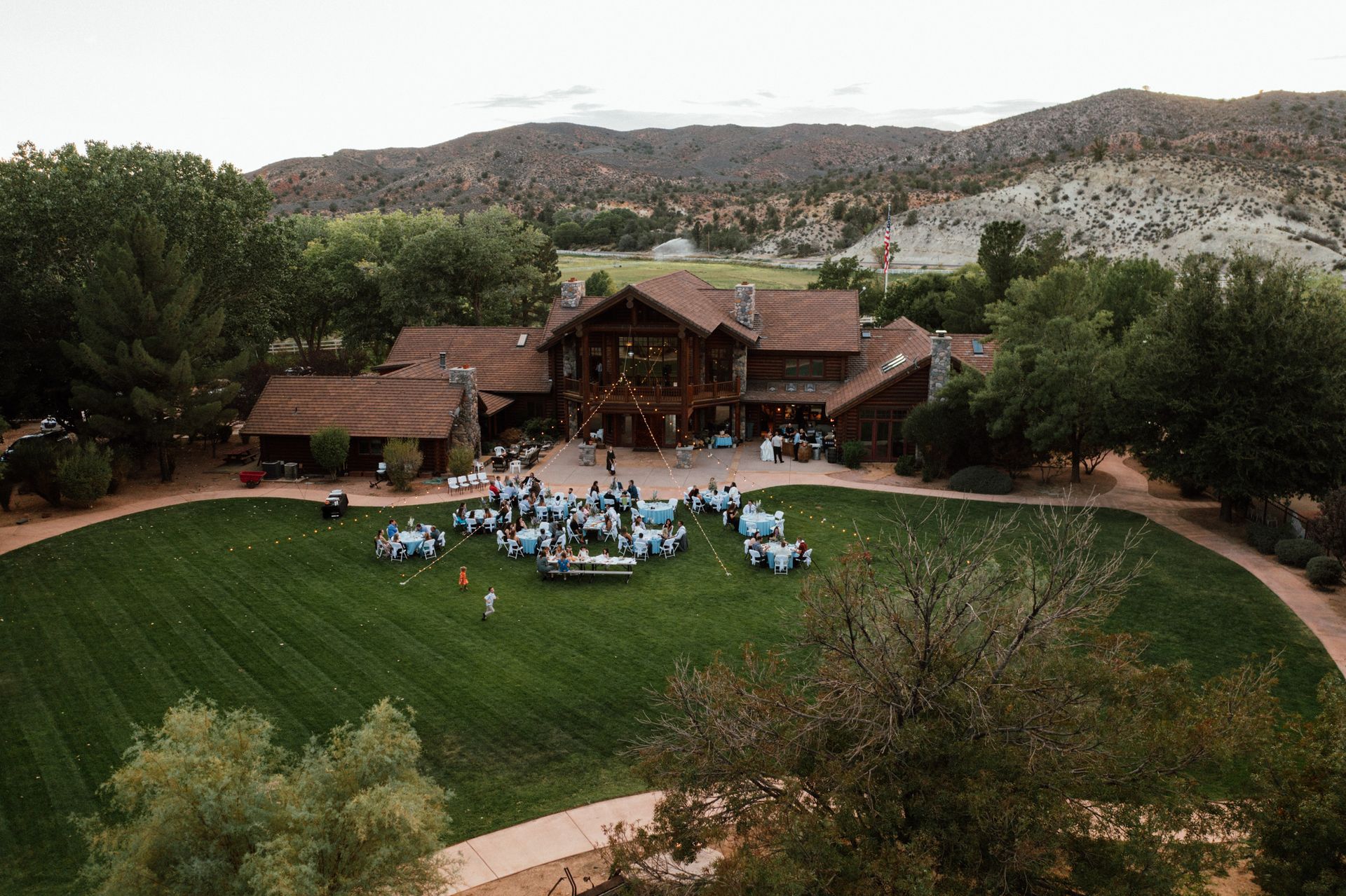 Outdoor gathering on a lawn in front of a rustic lodge, with hills in the background