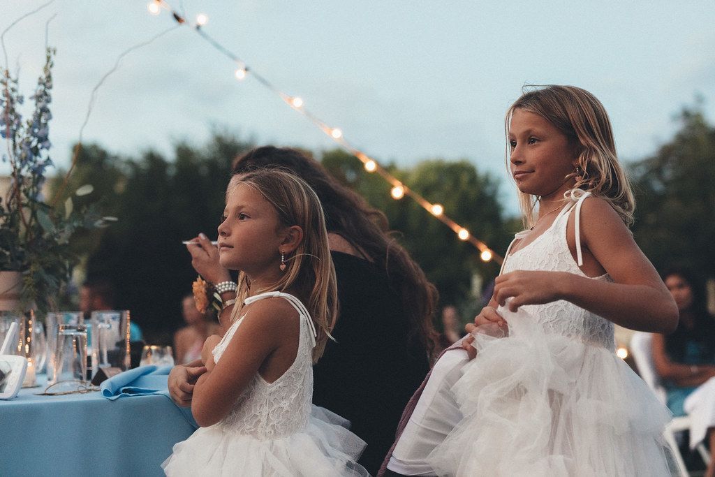 Two children in white dresses dancing at an outdoor evening celebration with string lights