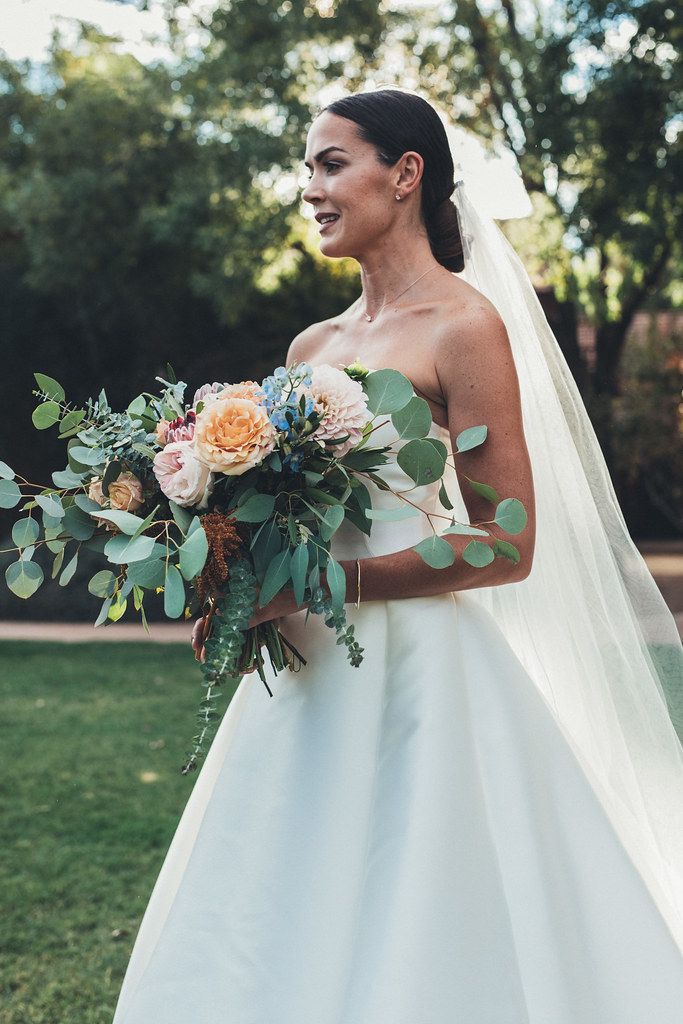 Bride in a white gown holding a pastel bouquet outdoors with a veil flowing behind her