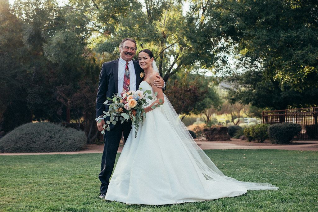 Bride and groom posing outdoors on a lawn, holding a bouquet, with trees in the background
