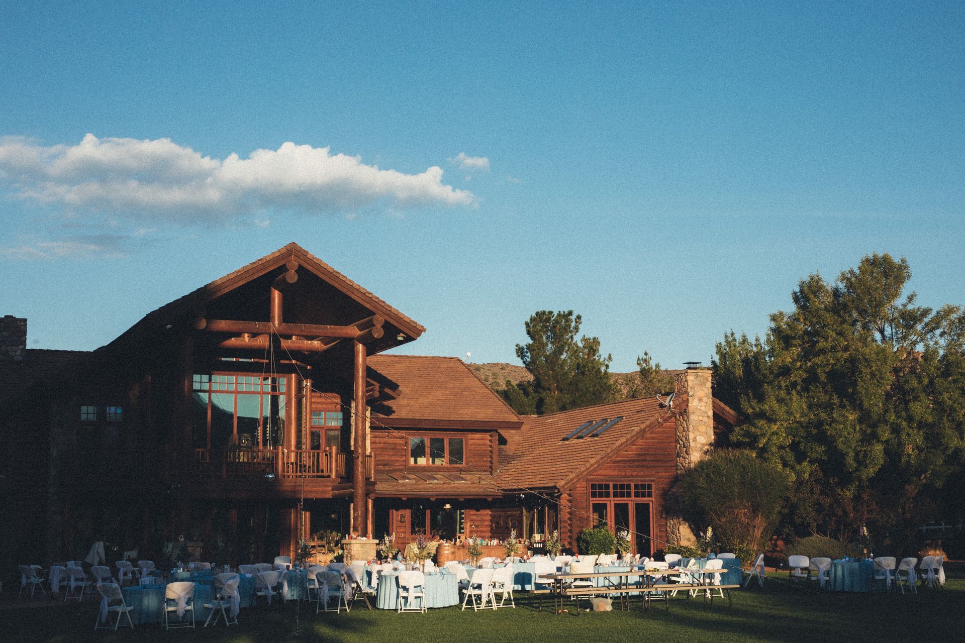 Rustic lodge with red roof and outdoor patio under a blue sky, surrounded by trees.