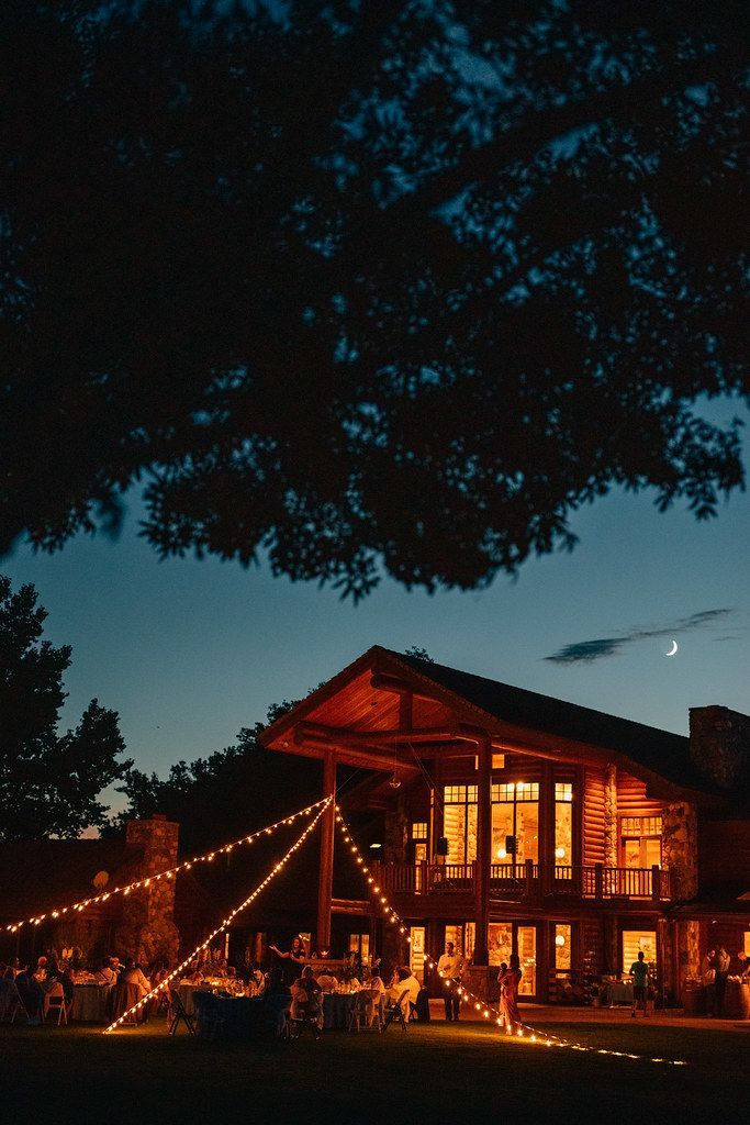 Lit log cabin glowing warmly at dusk under a dark tree canopy, with string lights and a fire pit outside