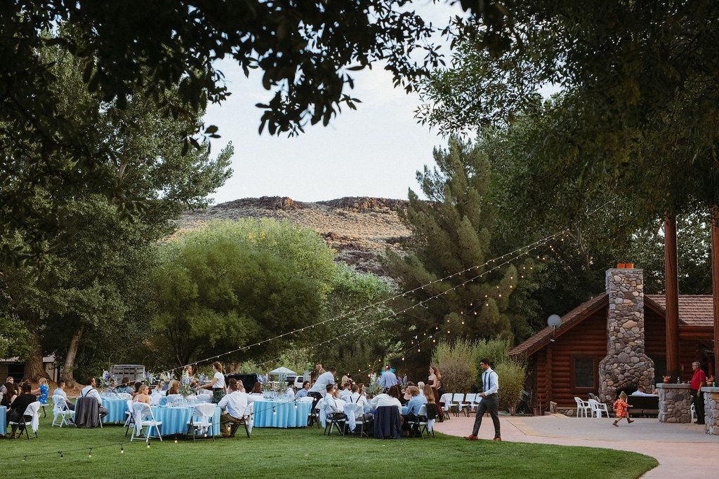 Outdoor wedding reception with guests seated on a lawn beside a rustic log cabin and stone chimney