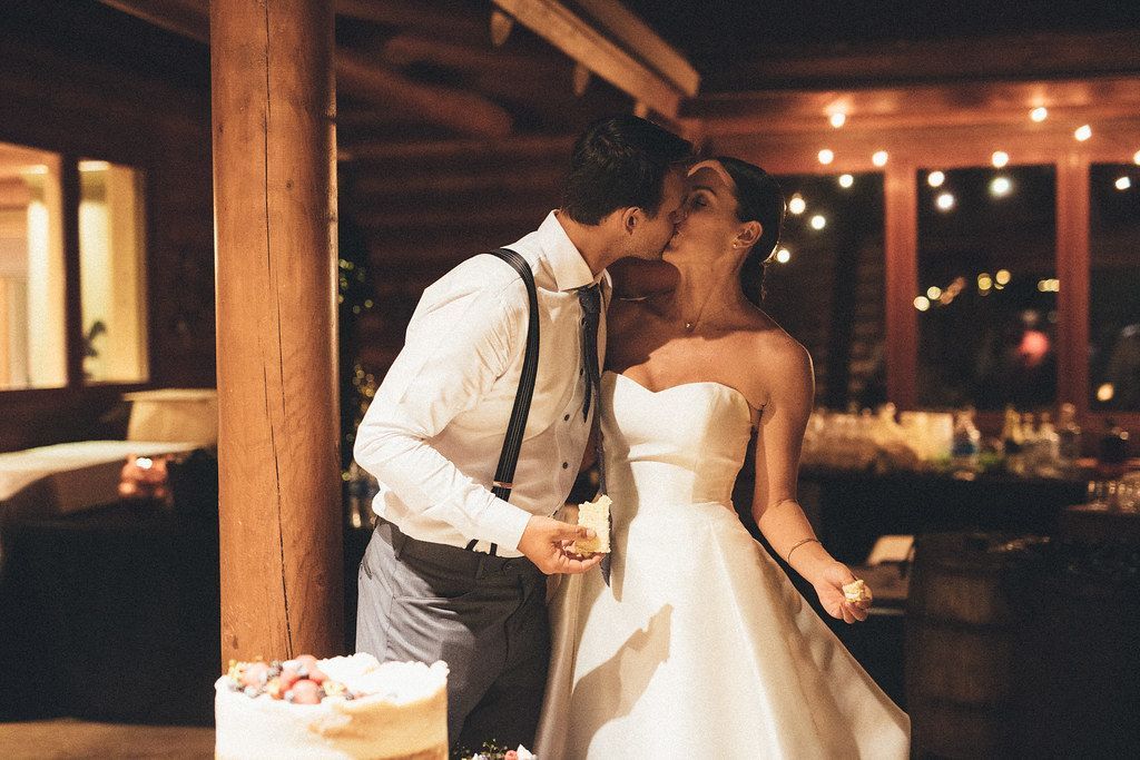 Couple kissing at a rustic indoor wedding reception, with string lights and a cake on the table