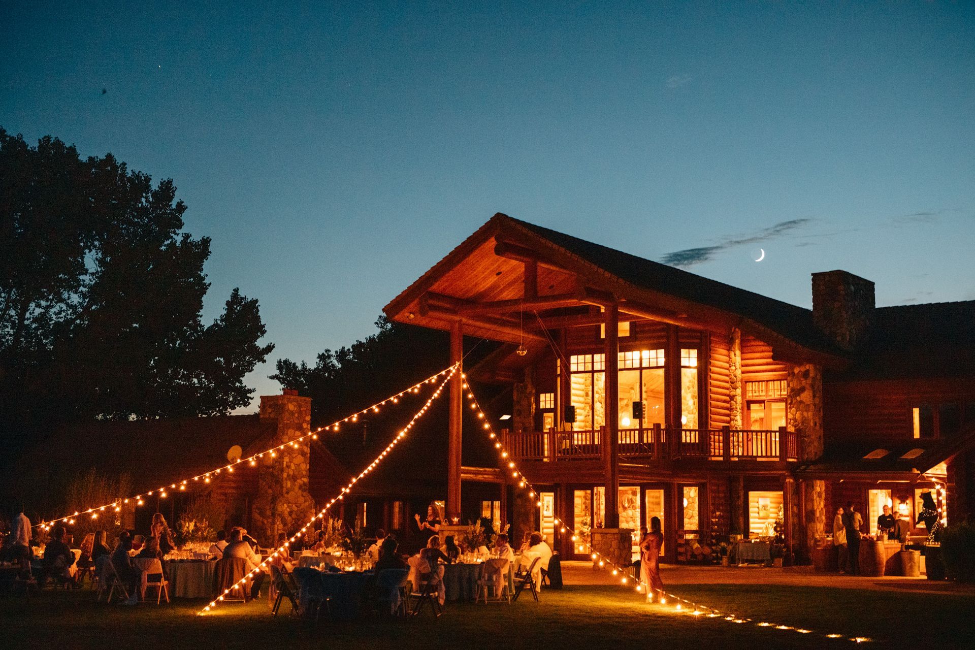 Lit wooden lodge lit by string lights at dusk, with people gathered outside on the lawn