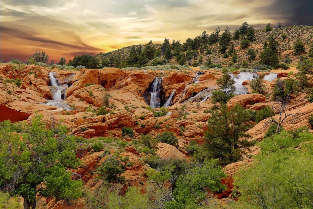 Red rock canyon with small waterfalls, green shrubs, and a cloudy sunset sky