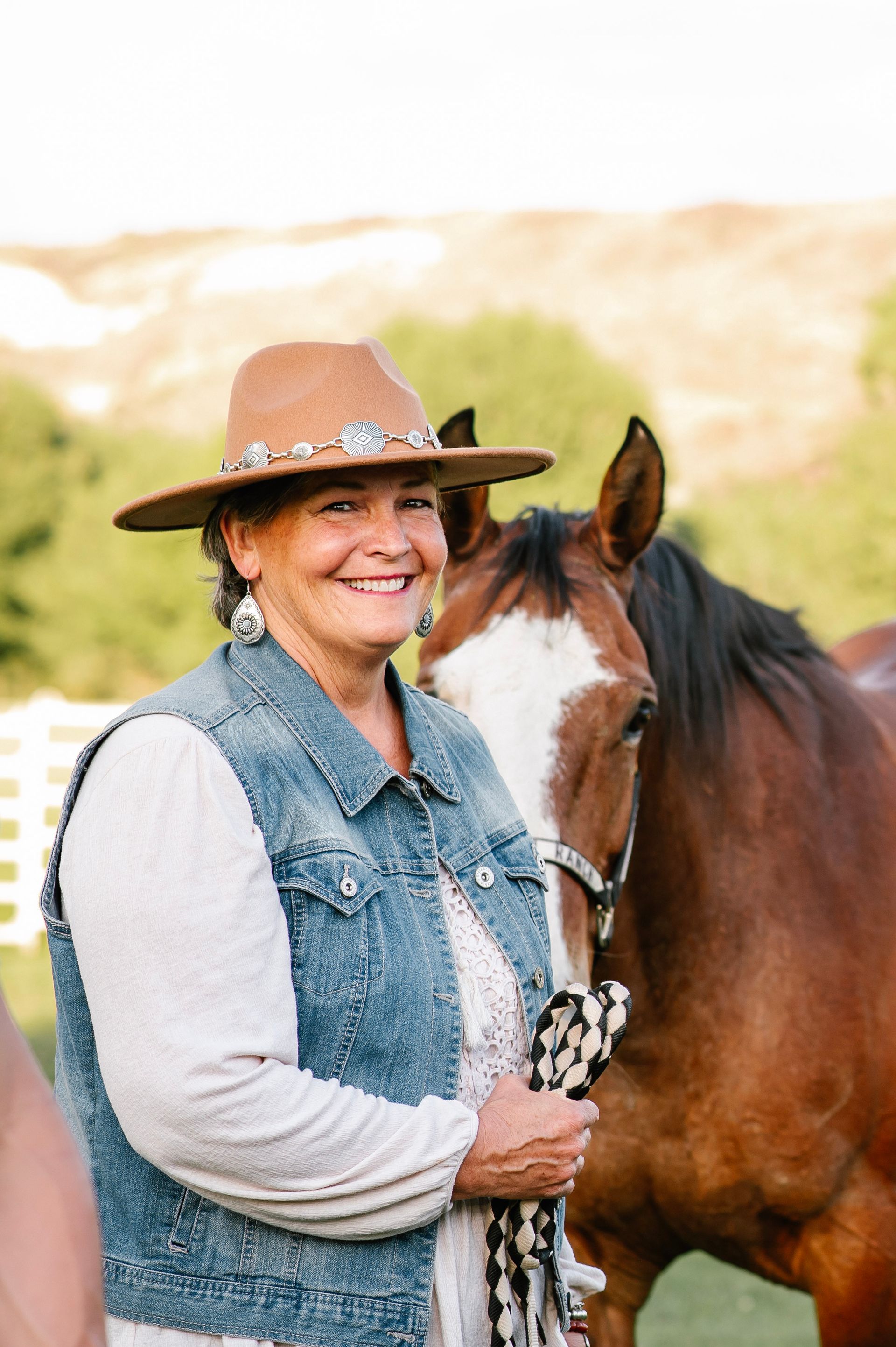 Smiling person in a brown hat stands beside a brown horse outdoors.