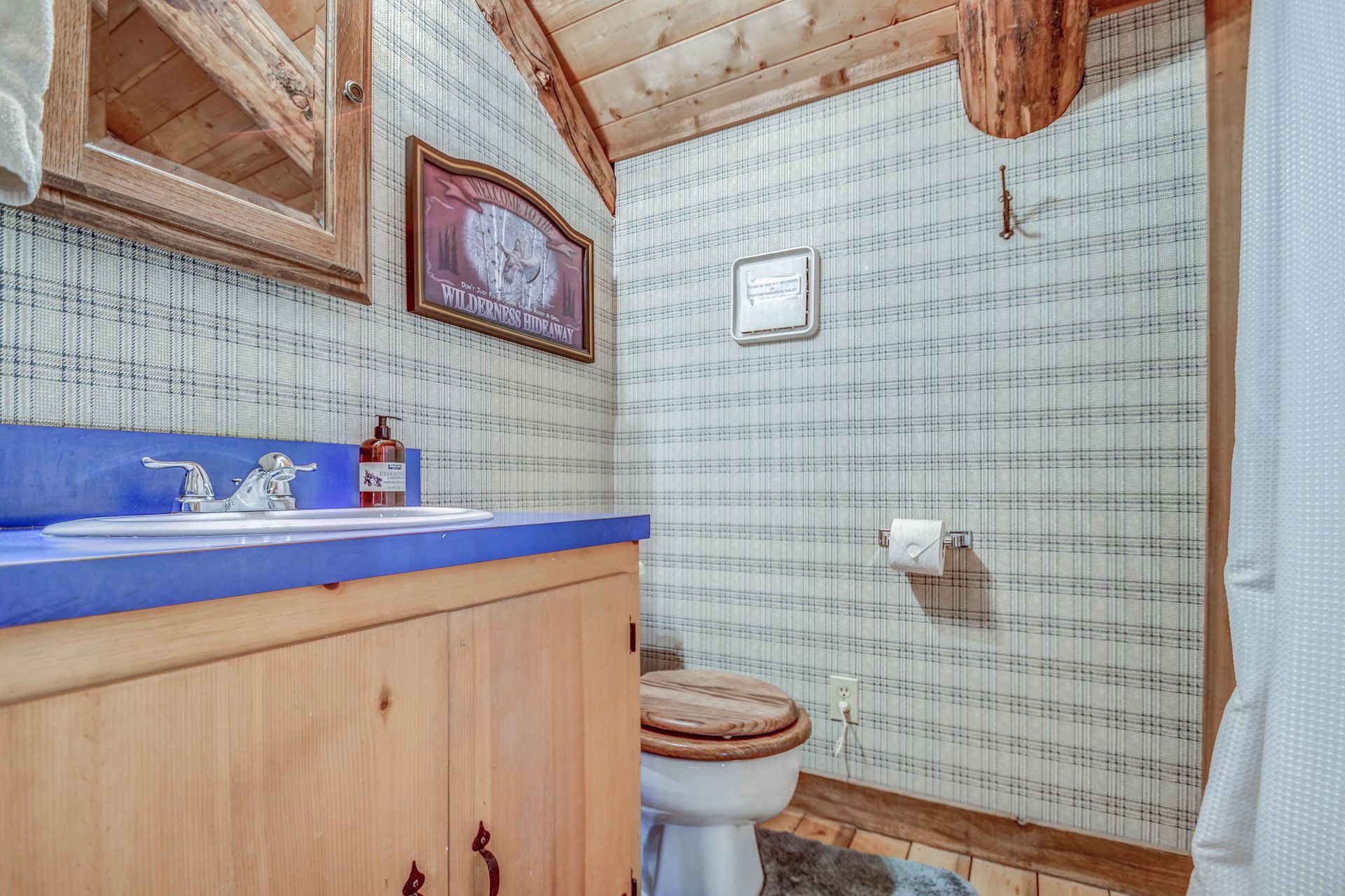 Small bathroom with wood vanity, blue countertop, toilet, patterned wallpaper, and shower curtain.