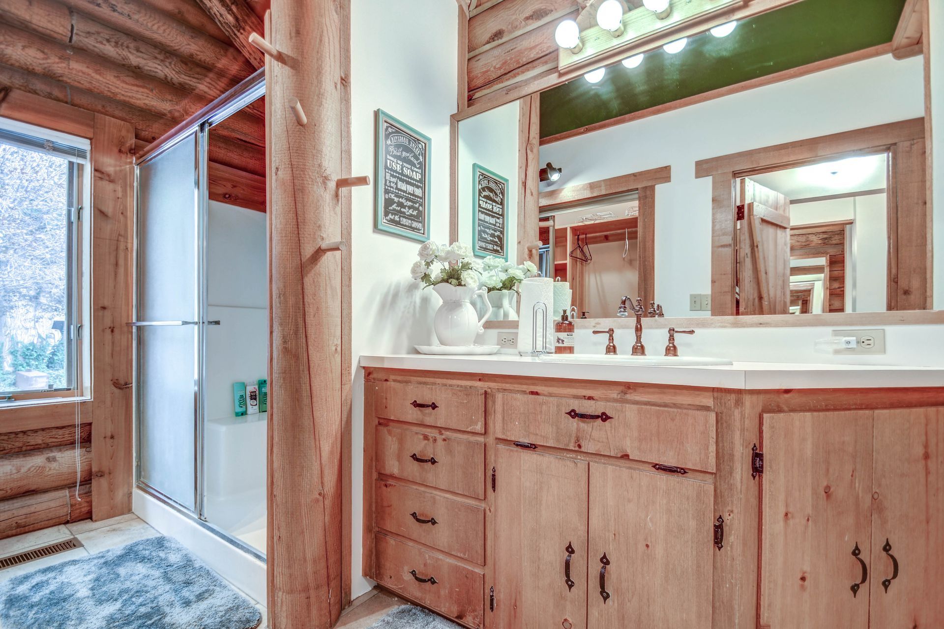 Wood-paneled bathroom with double vanity, large mirror, and a shower area with blue rug by a snowy window
