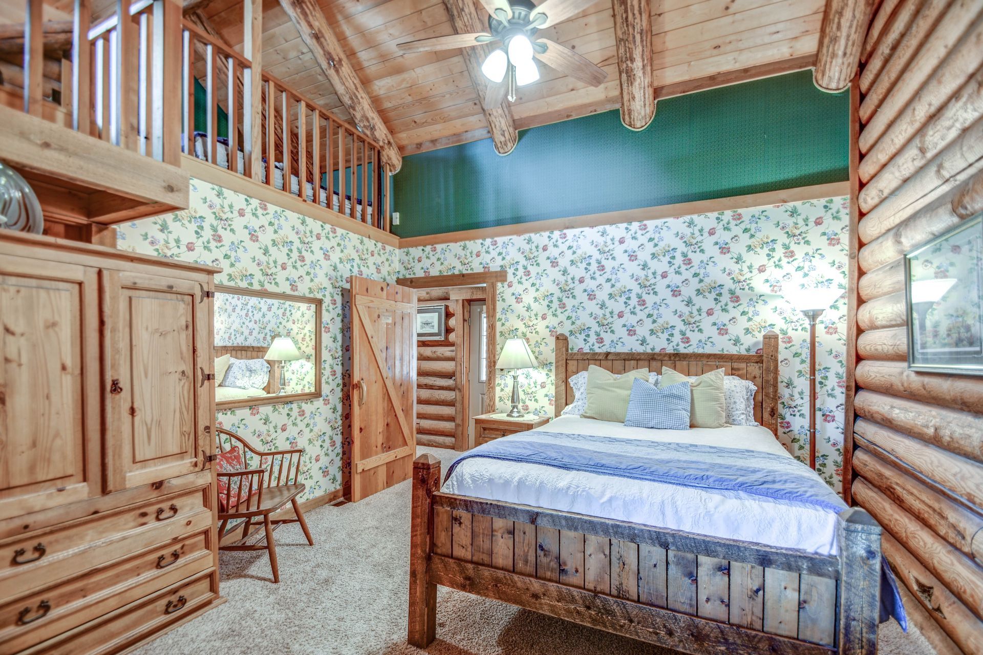 Rustic log cabin bedroom with wood bed, floral wallpaper, loft railing, and ceiling fan
