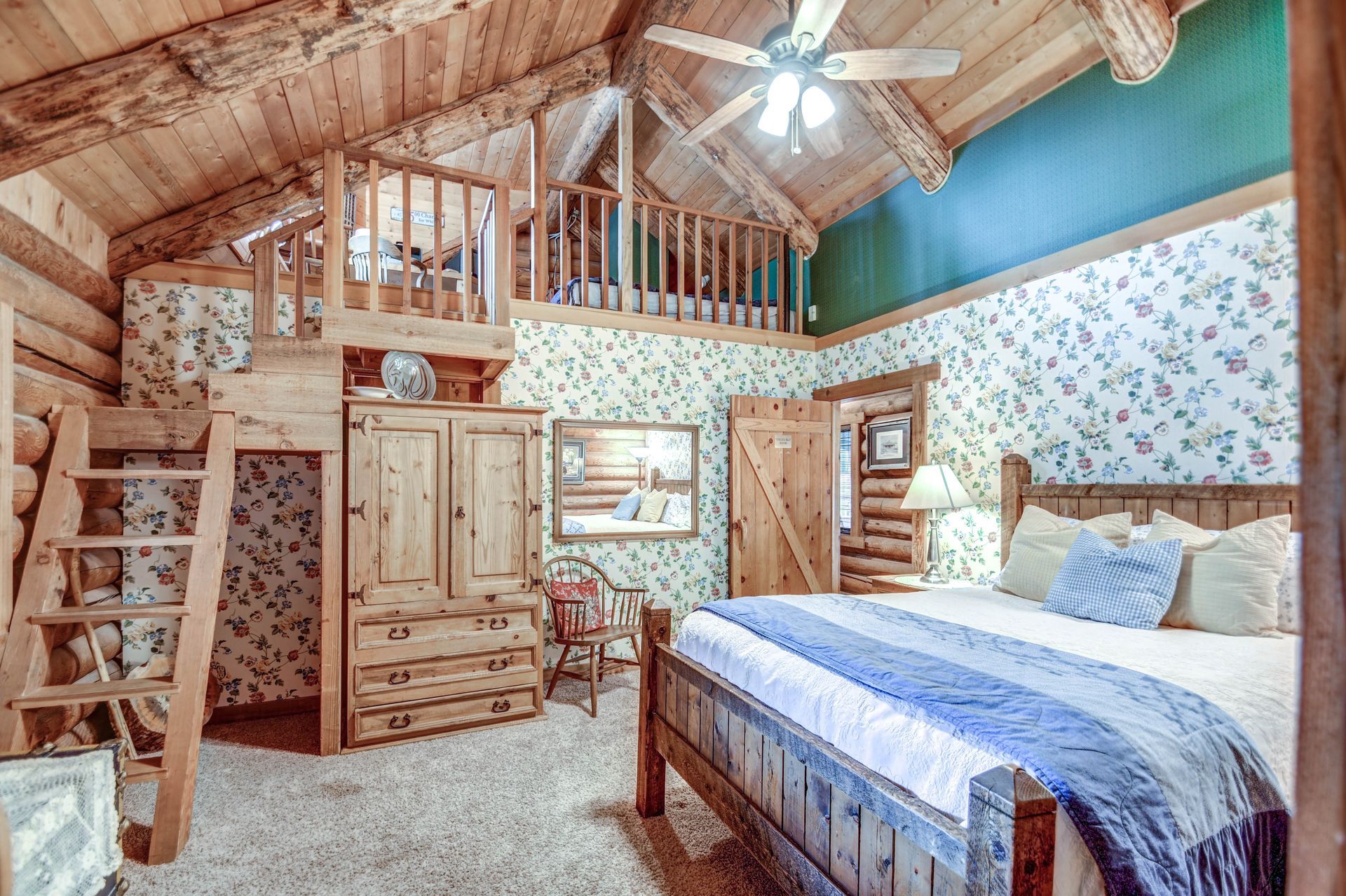 Cozy rustic loft bedroom with wooden walls, blue bedspread, and a ceiling fan under vaulted wood beams