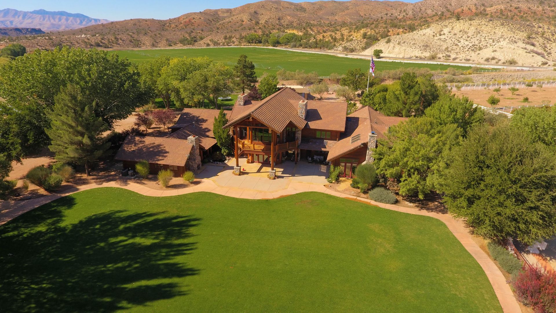 Aerial view of a large rustic house with a green lawn, trees, and desert hills in the background.