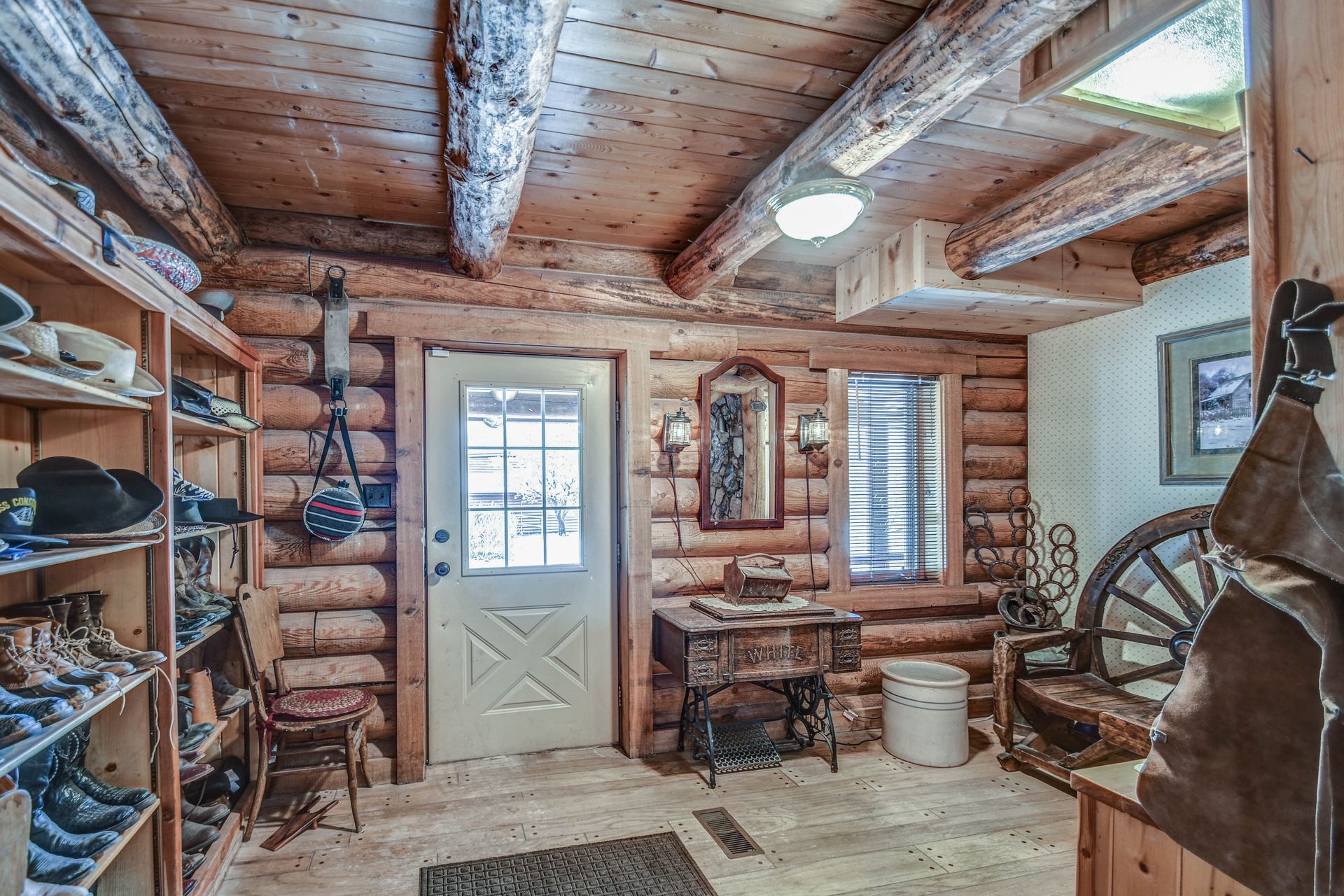 Rustic log cabin interior with wooden beams, small desk, shelves, door, and daylight through windows