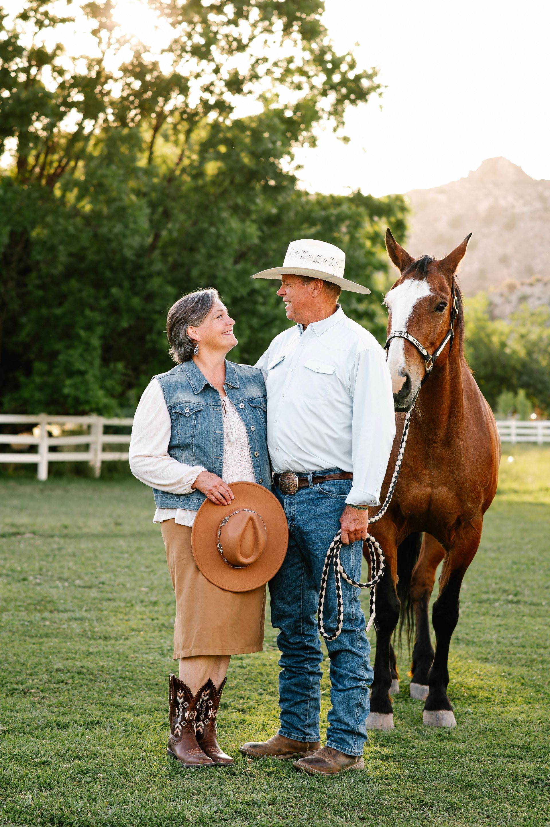 Two people standing with a horse in a grassy field at sunset, one holding a hat.
