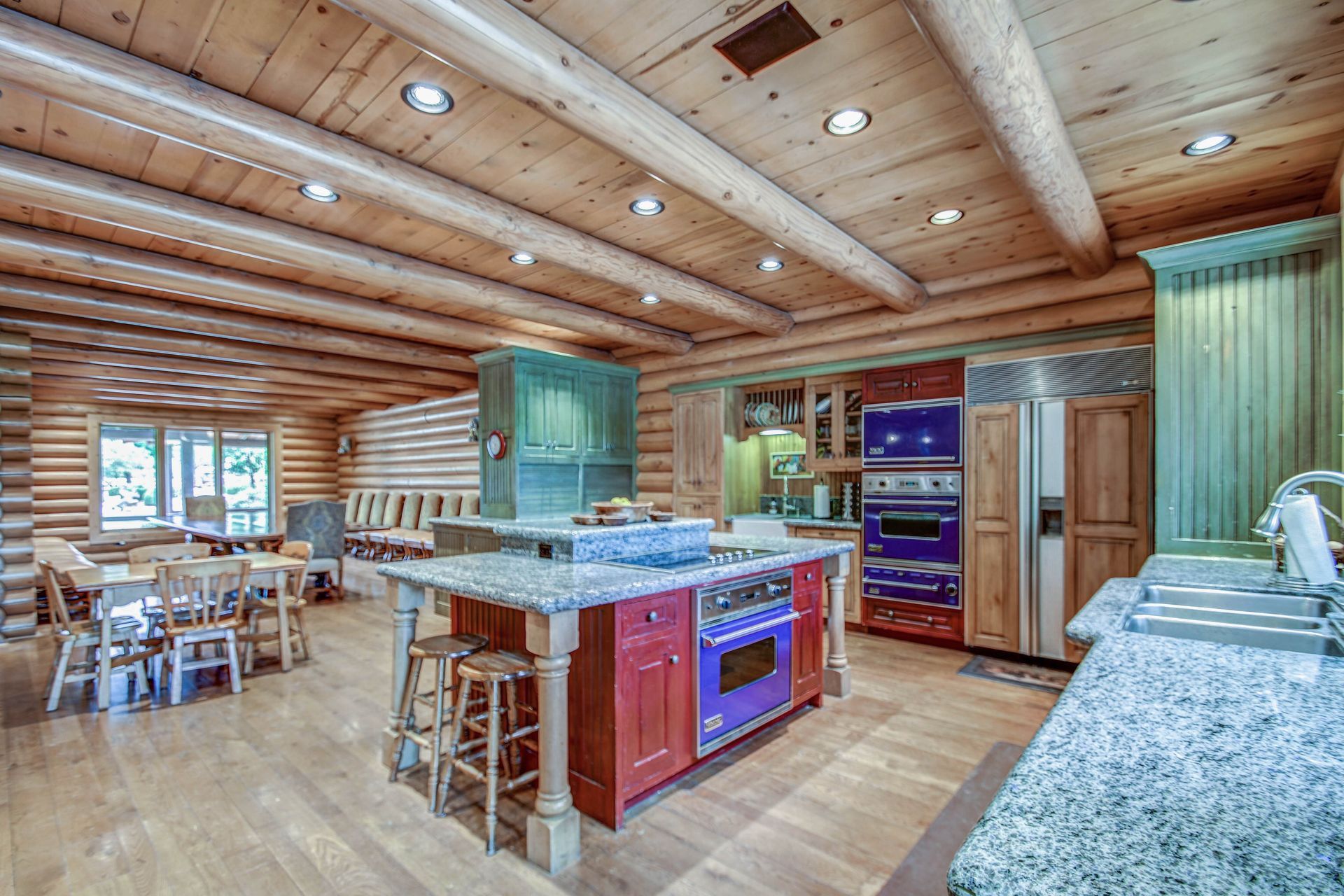 Rustic log cabin kitchen with green cabinets, granite counters, and a wood ceiling, open to a dining area