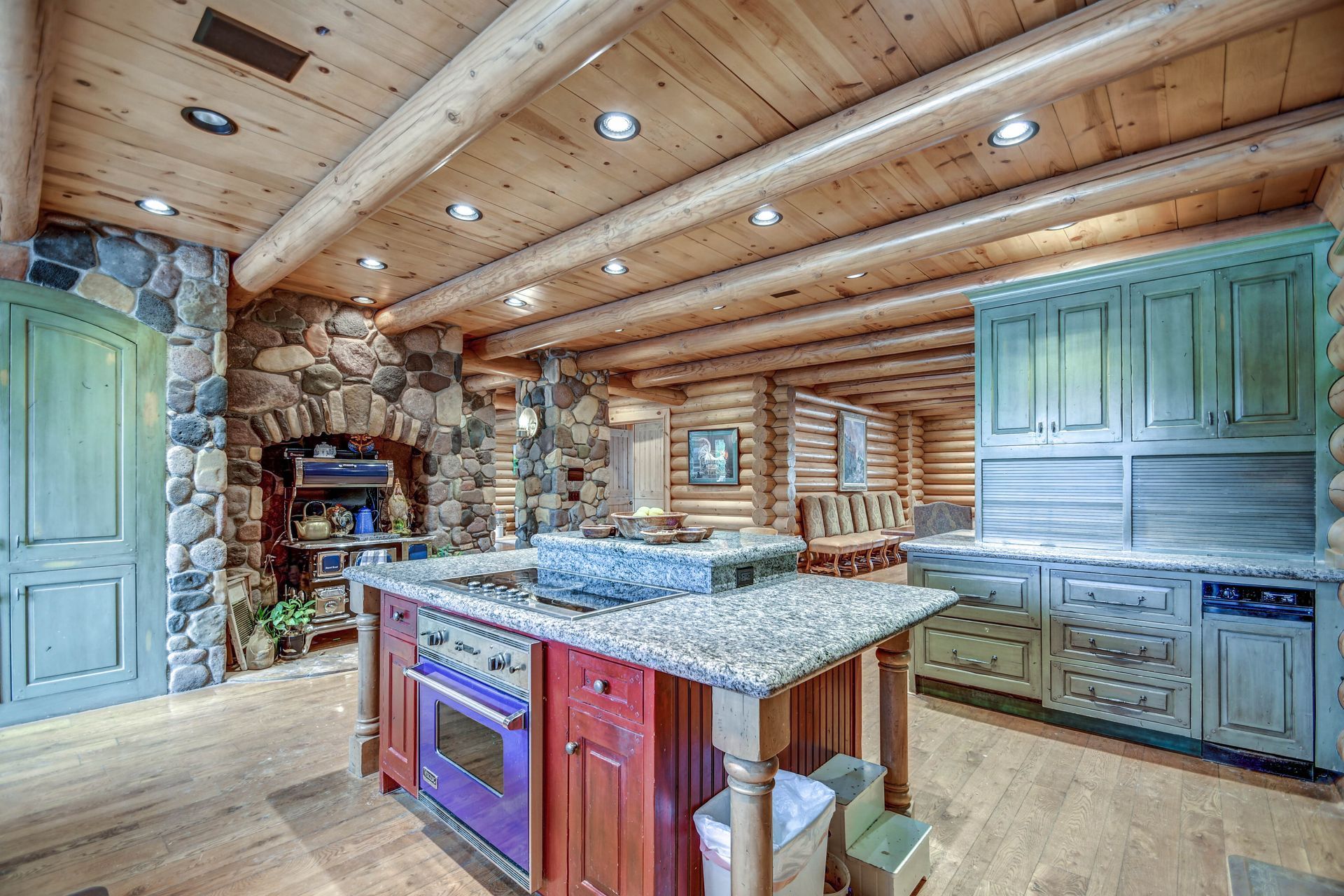 Rustic log cabin kitchen with granite island, teal cabinets, stone fireplace, and wood beam ceiling.