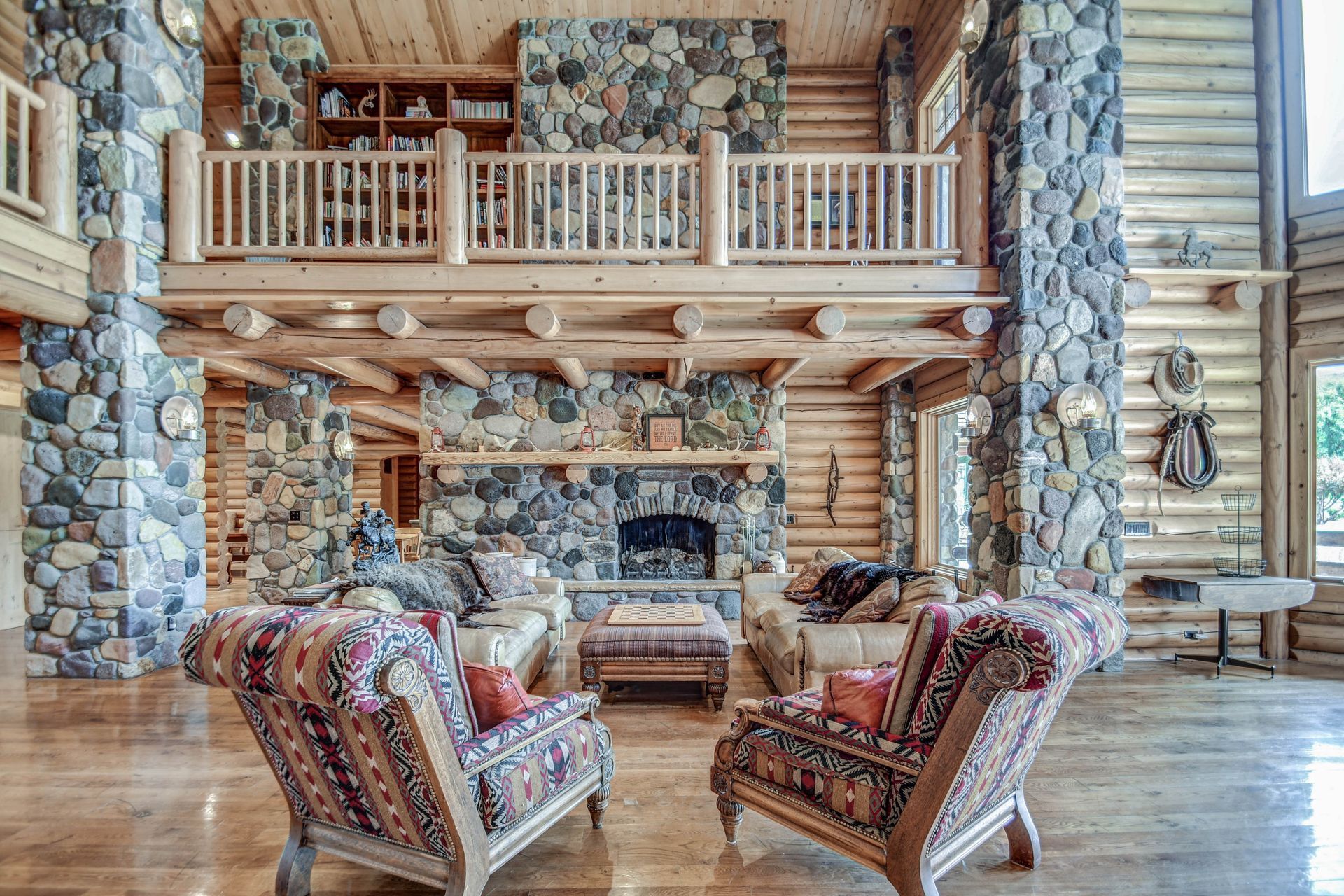 Rustic lodge living room with stone fireplace, wood balcony, and patterned armchairs on hardwood floor