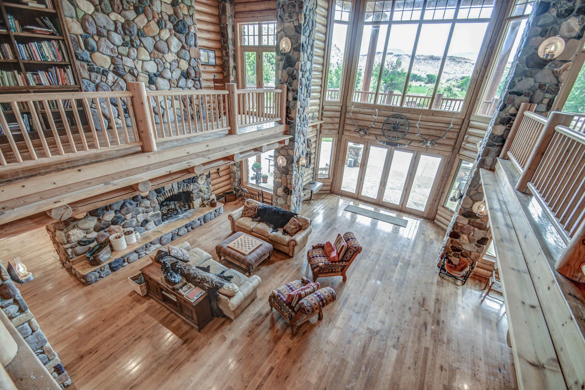 Rustic log cabin great room with stone fireplace, wood floors, and large windows overlooking a loft