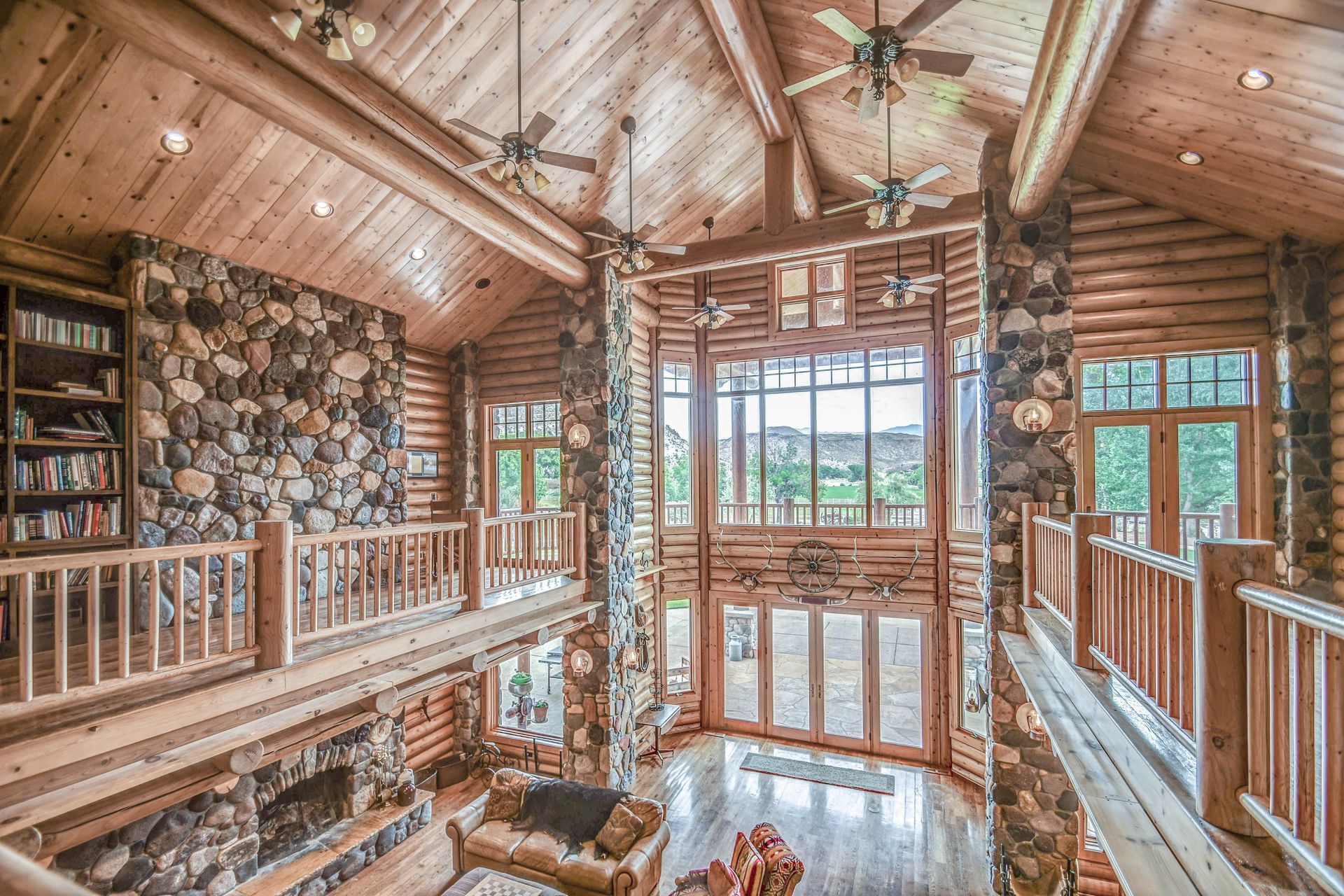Rustic log cabin living room with stone fireplace, wood ceiling, balcony, and large windows