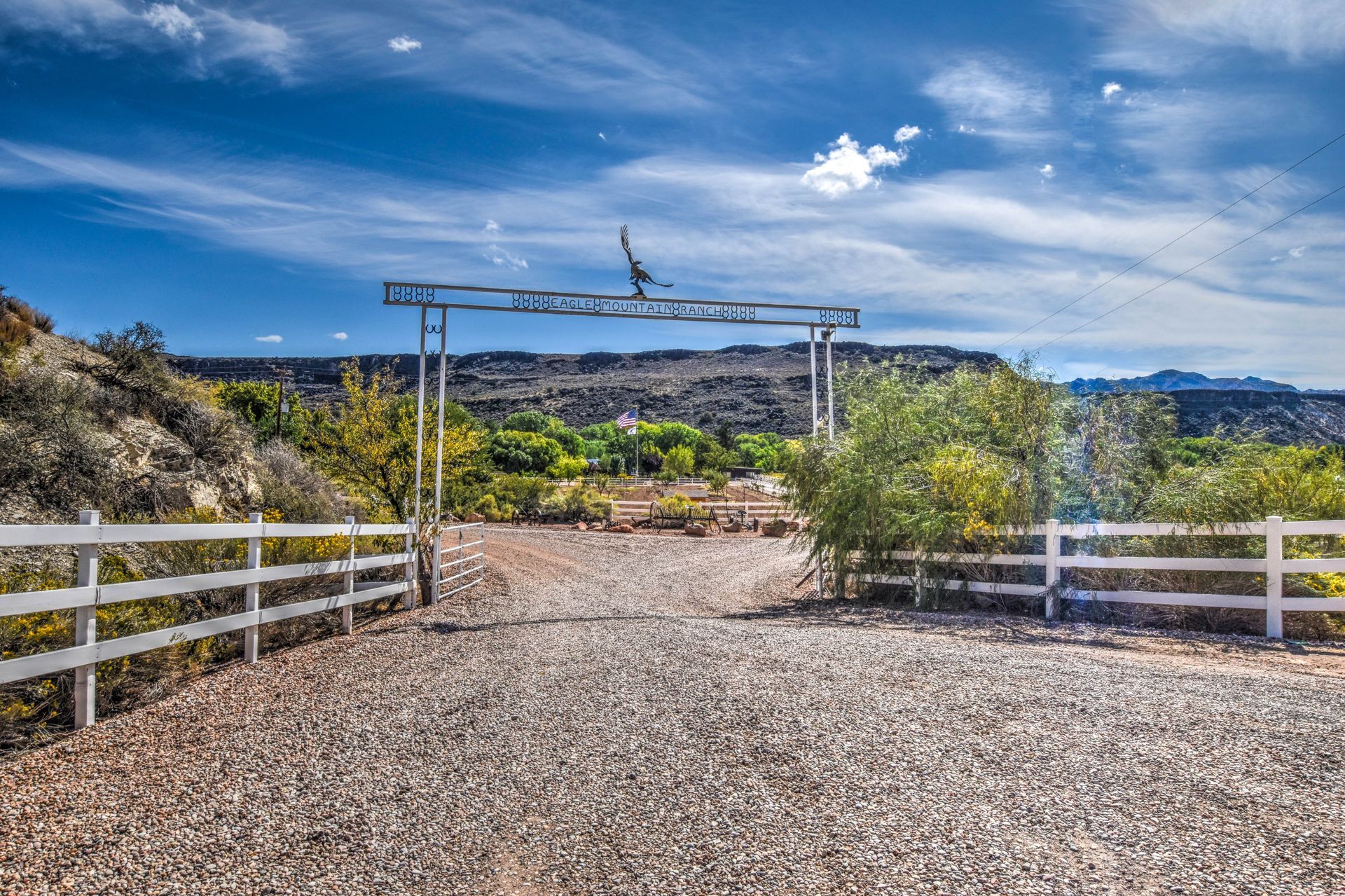 Gravel driveway gate with white fences, desert plants, and distant hills under a blue sky